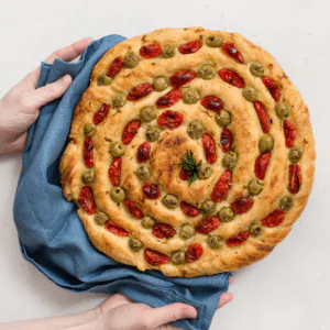 Hands holding a round focaccia decorated with a spiral of cherry tomatoes and green olives