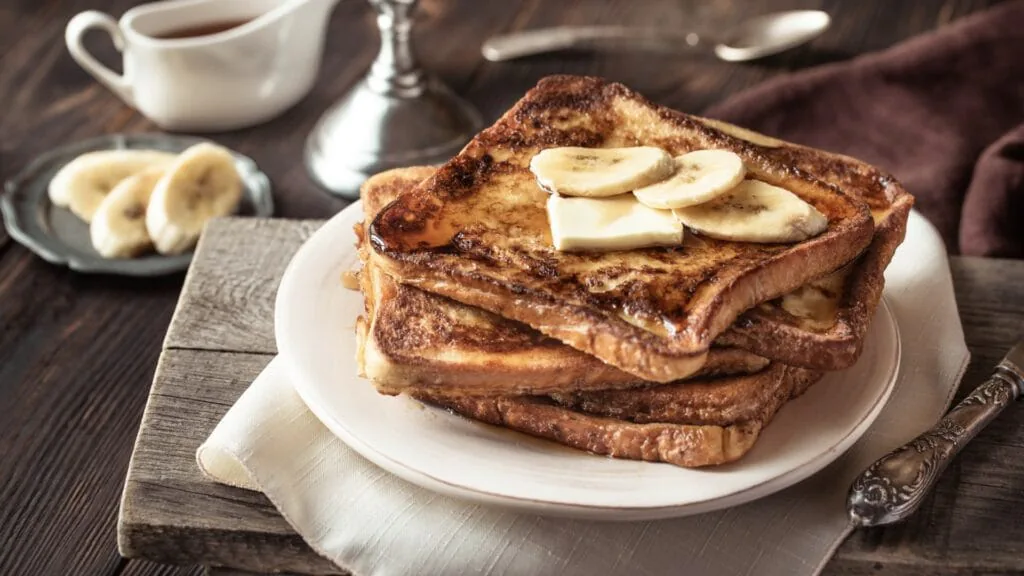 Stack of French toast topped with banana slices and butter, with a jug of syrup alongside