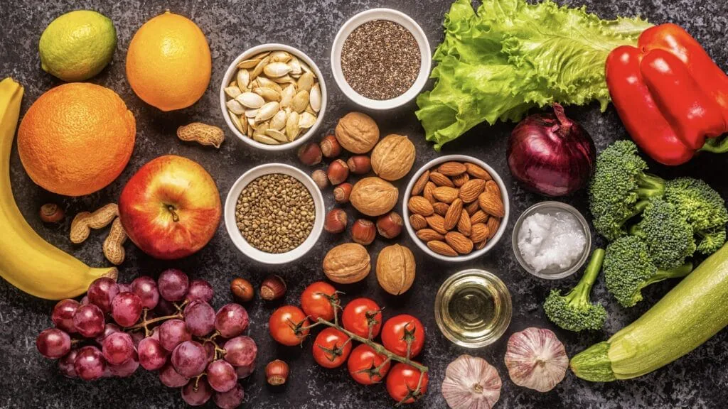 Overhead spread of fresh vegetables, fruit, nuts and seeds on a dark stone surface