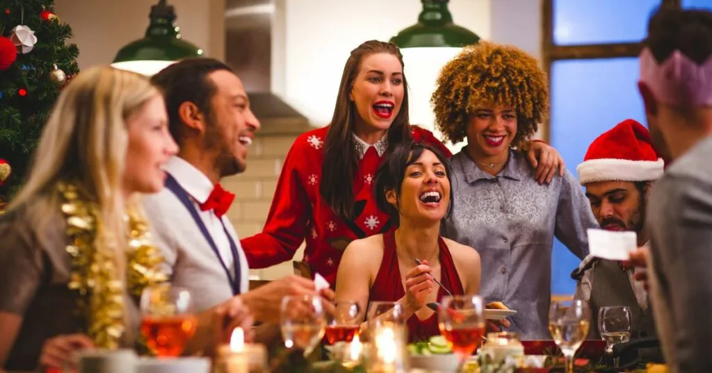 Group of coworkers laughing around a festive dinner table with drinks and a Christmas tree