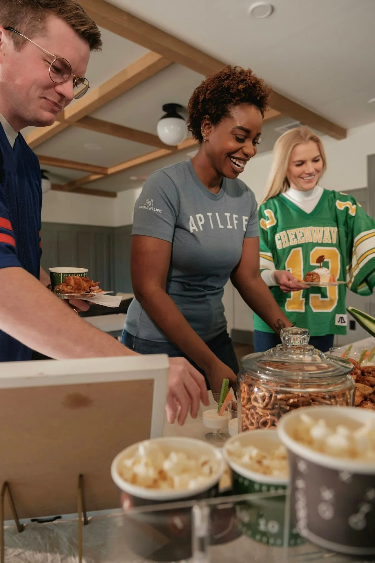 People serving themselves popcorn and pretzels at a game-day snack table, one wearing a football jersey