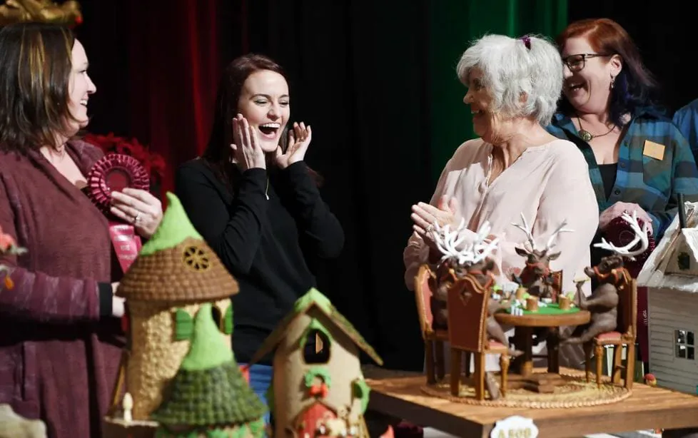 Four coworkers laughing beside elaborately decorated gingerbread houses displayed on a table