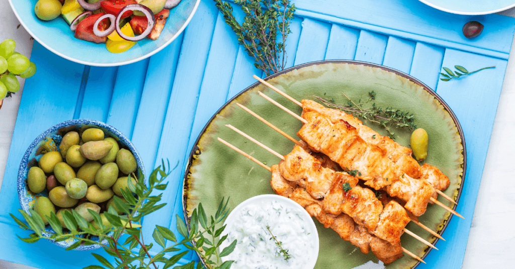 Grilled chicken skewers on a green plate with tzatziki, olives and a Greek salad