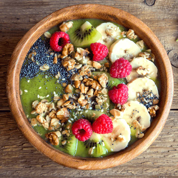 Green smoothie bowl topped with raspberries, banana, kiwi, walnuts and chia seeds in a wooden bowl