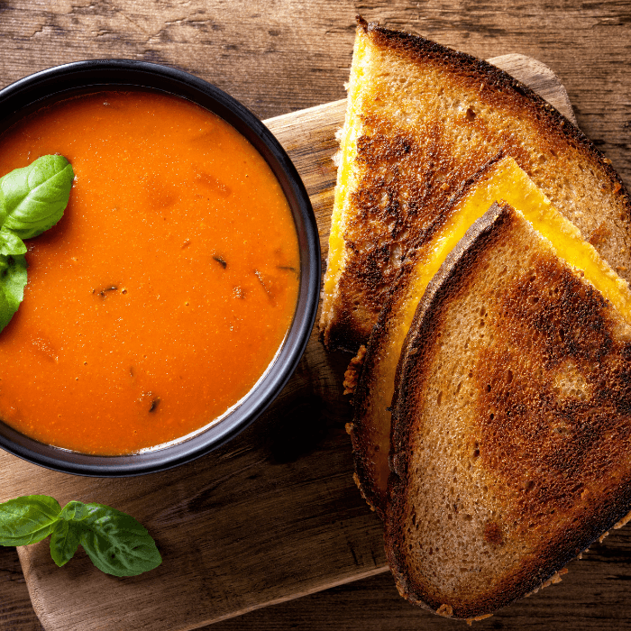 Grilled cheese sandwich cut in half on a wooden board with a bowl of tomato soup and basil