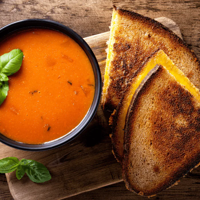 Grilled cheese sandwich cut in half on a wooden board with a bowl of tomato soup and basil