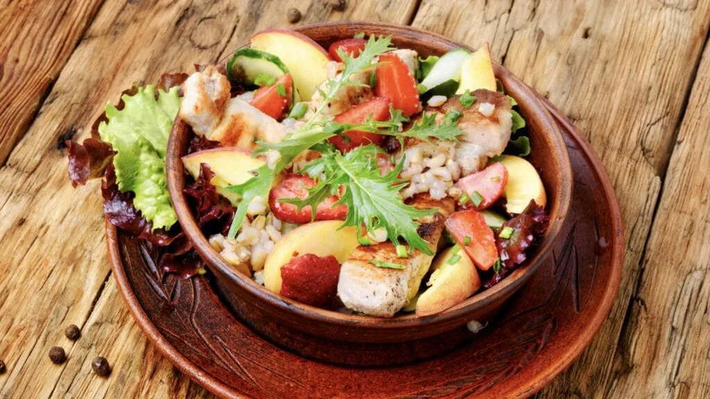 Warm grain salad of grilled chicken, barley, strawberries and peach slices with arugula in a wooden bowl
