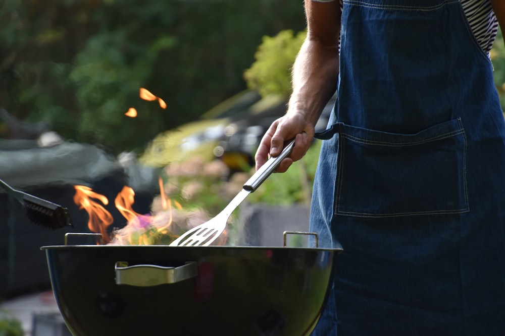 Person in a denim apron turning food with tongs over a flaming charcoal grill outdoors