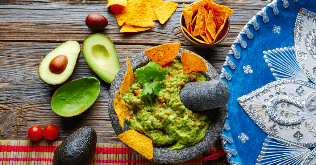 Guacamole in a stone molcajete with tortilla chips, halved avocados and a blue sombrero