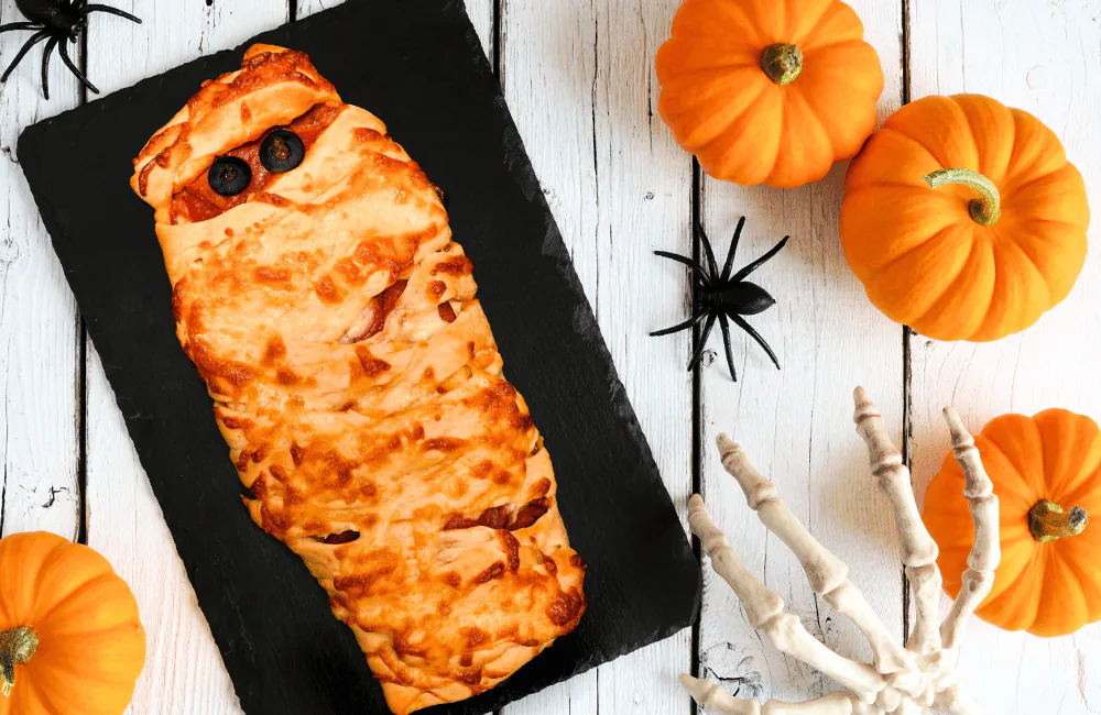 Mummy-shaped calzone on a slate board with mini pumpkins, plastic spiders and a skeleton hand