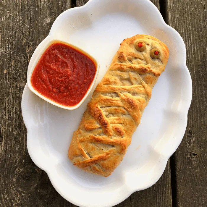 Mummy-shaped pastry roll with olive eyes on a white plate beside a bowl of marinara sauce