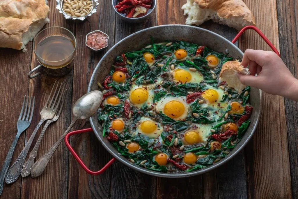 Hand dipping crusty bread into a pan of eggs baked with spinach and sun-dried tomatoes