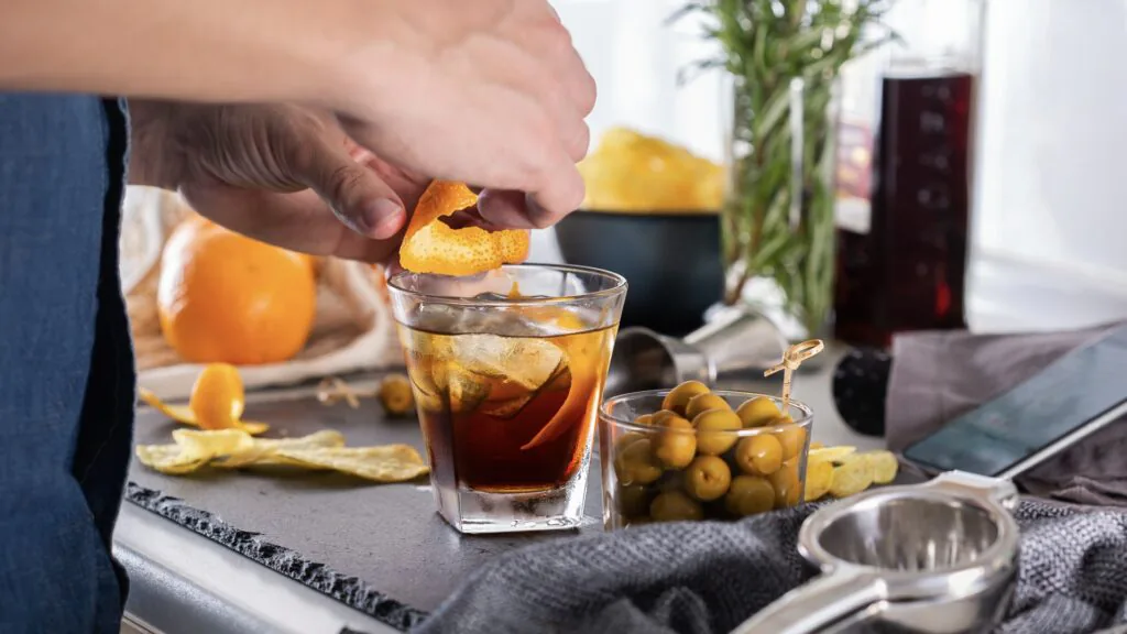Hand twisting an orange peel over a dark cocktail in a rocks glass beside a bowl of olives