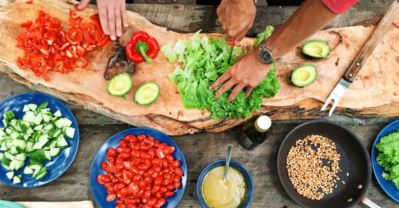 Overhead view of hands chopping peppers and lettuce on a rustic wooden board ringed by bowls of vegetables