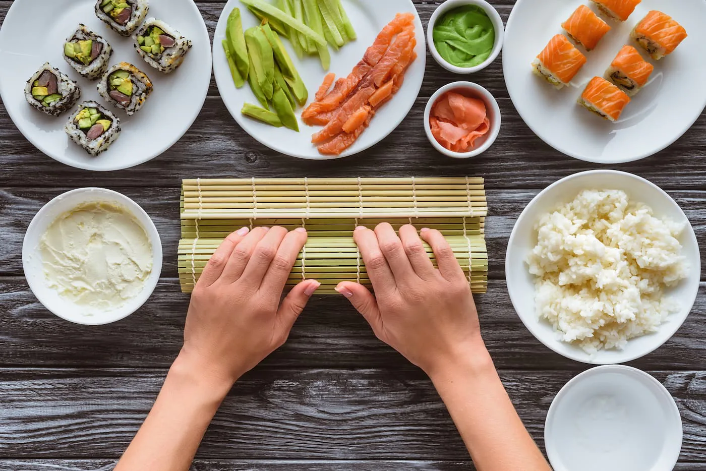 Overhead view of hands on a bamboo mat surrounded by salmon, cucumber, rice and finished sushi rolls
