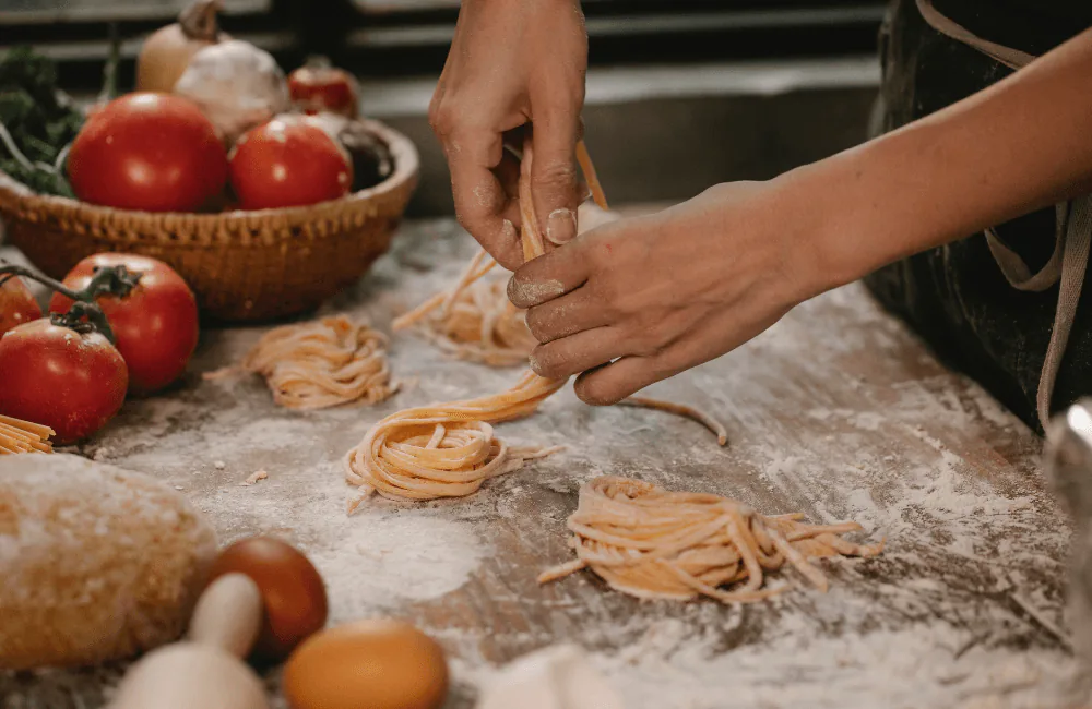 Hands shaping nests of fresh pasta on a floured counter with tomatoes and eggs nearby