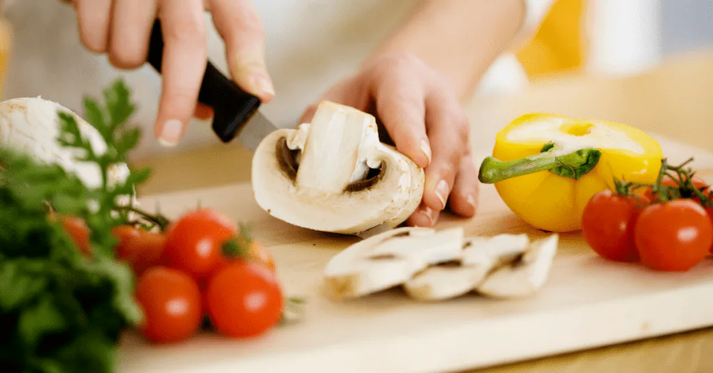 Hands slicing a white mushroom on a wooden board beside cherry tomatoes, parsley and a yellow pepper