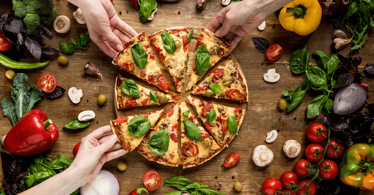 Hands reaching for slices of a basil and tomato pizza surrounded by fresh vegetables and mushrooms