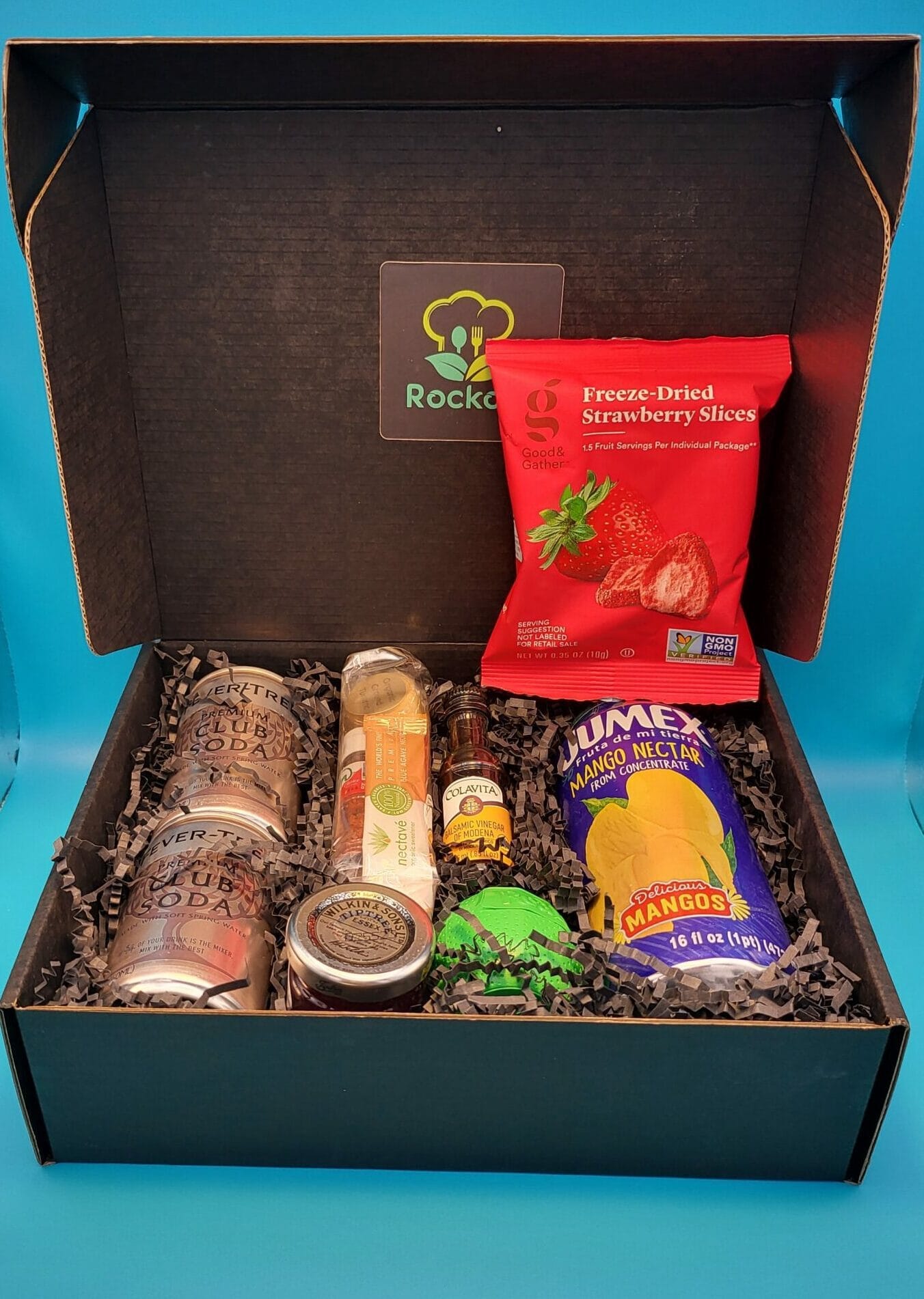 Open happy hour kit box with soda cans, mango nectar, bitters and freeze-dried strawberry slices