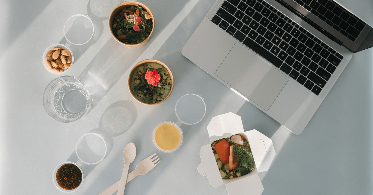 Overhead of two grain bowls with greens and edible flowers, juice and a takeout box beside a laptop
