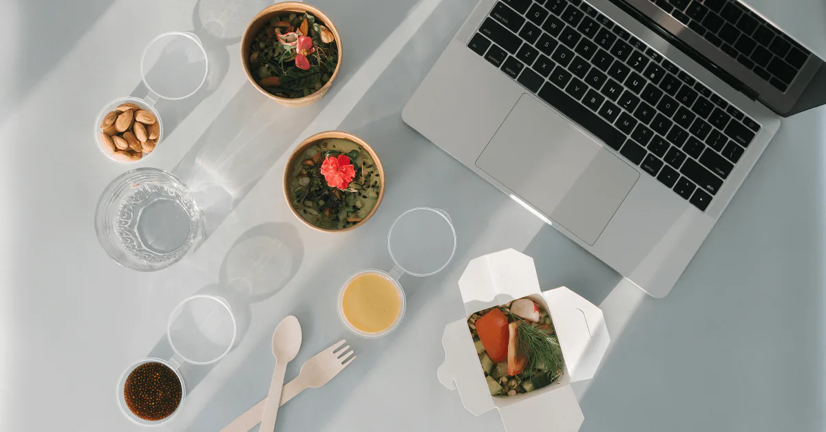Overhead of two grain bowls with greens and edible flowers, juice and a takeout box beside a laptop