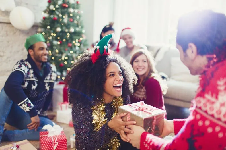 Laughing woman receives a wrapped gift from a colleague at a holiday party by the Christmas tree