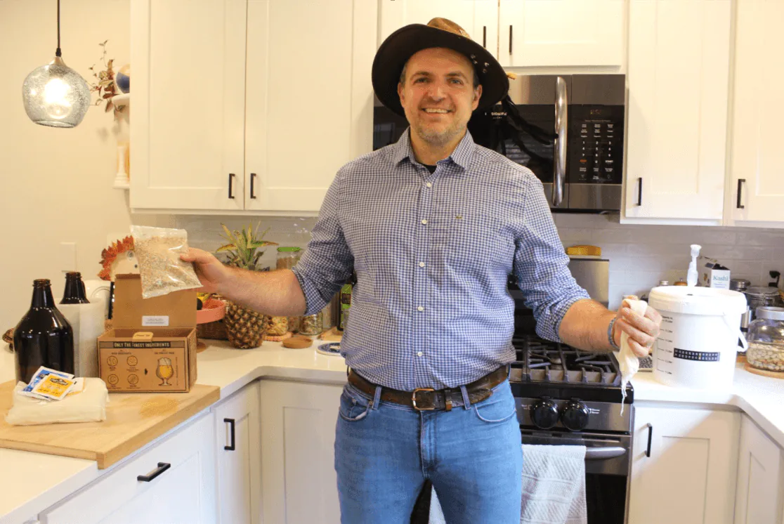 Host in a home kitchen holding a bag of malt beside a beer brewing kit, fermenting bucket and growlers