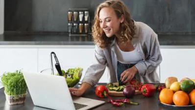 Smiling home cook following a laptop in her kitchen while chopping fresh vegetables and peppers
