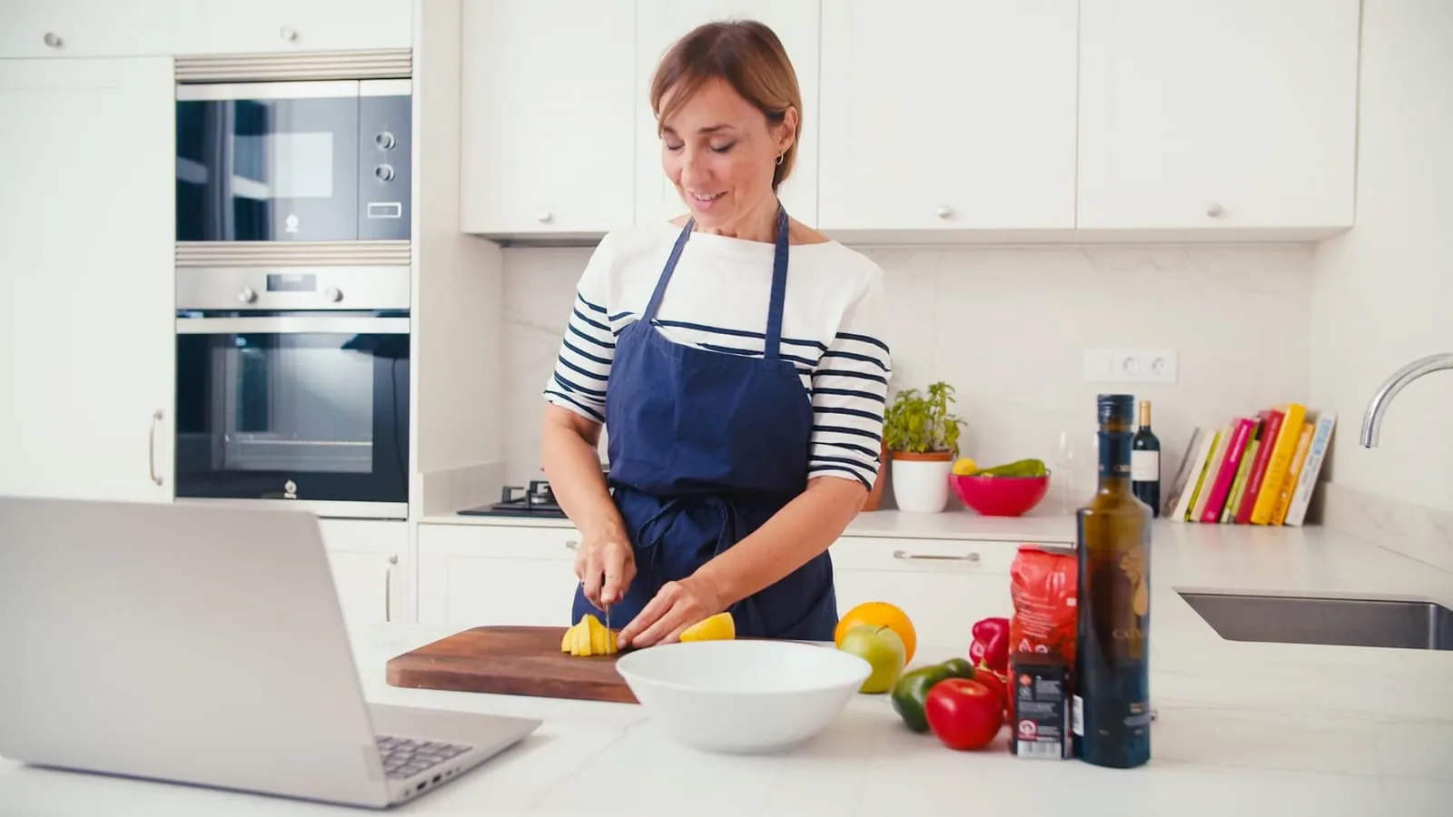Woman in a navy apron slicing a lemon in a white kitchen with an open laptop on the counter