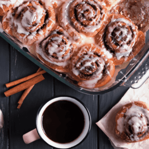 Overhead pan of iced cinnamon rolls with cinnamon sticks and a mug of black coffee
