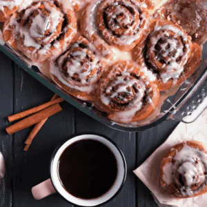 Overhead pan of iced cinnamon rolls with cinnamon sticks and a mug of black coffee