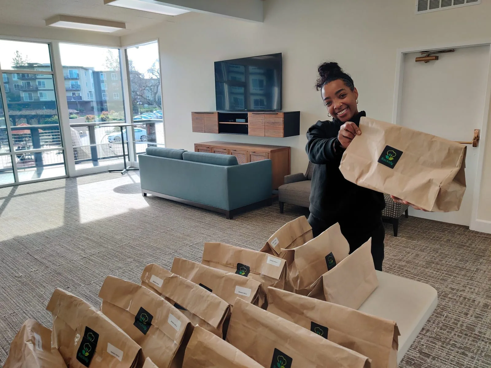Event host grinning as she holds up paper ingredient kits beside a table stacked with more bags