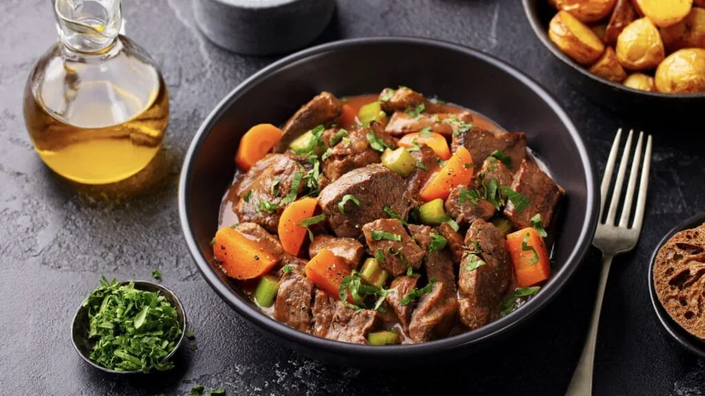 Bowl of Irish beef stew with carrots, celery and parsley beside olive oil and roast potatoes