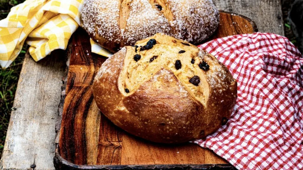 Two round loaves of Irish soda bread, one studded with raisins, on a wooden board with checked cloths