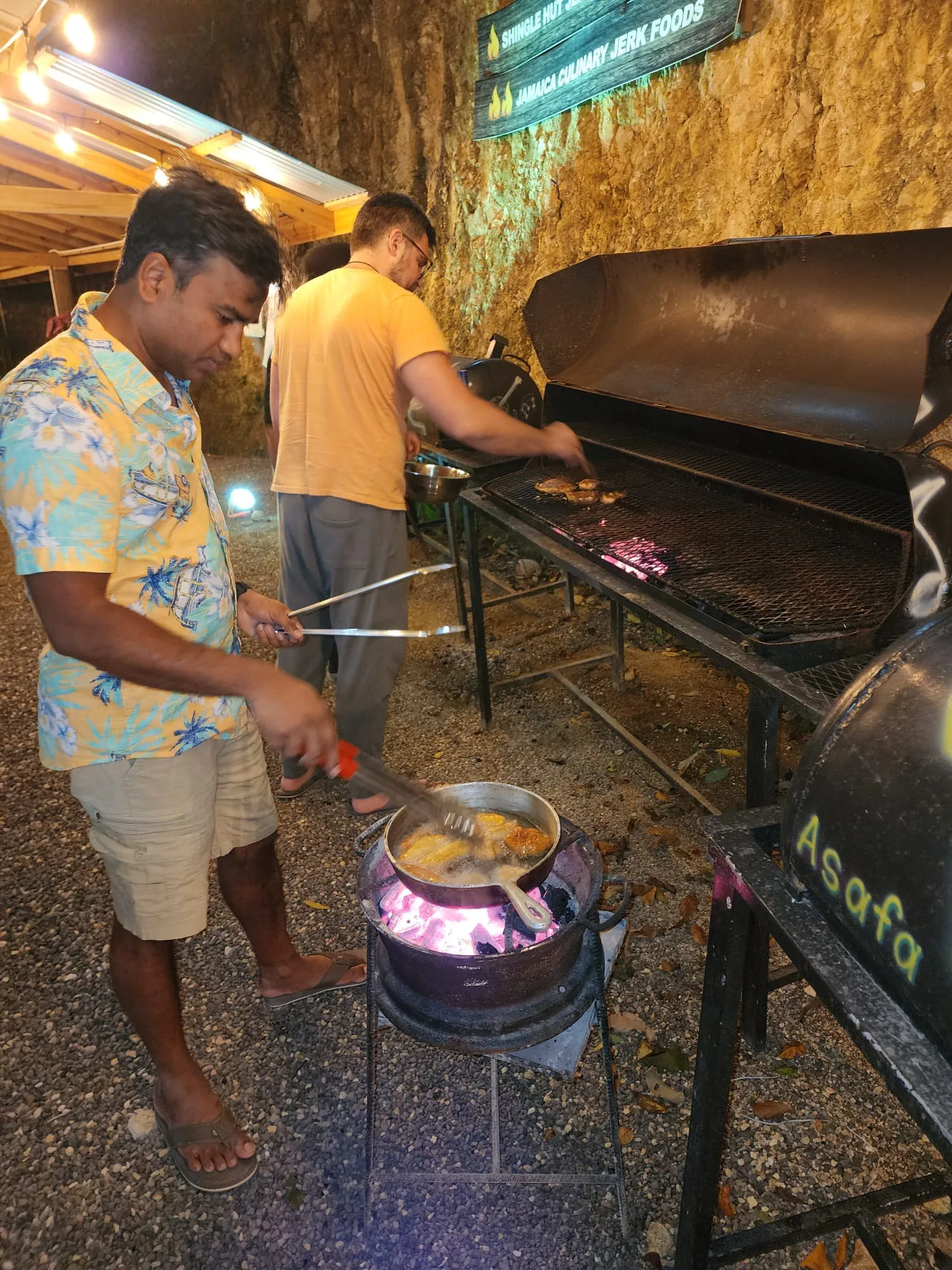 Cooks grilling jerk chicken and frying festival dough at an open-air Jamaican jerk stand at night