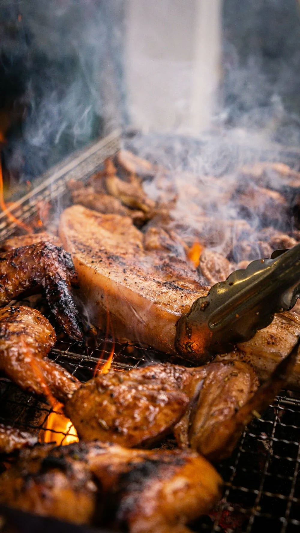 Jerk chicken pieces smoking over open flames as tongs turn one on the grill