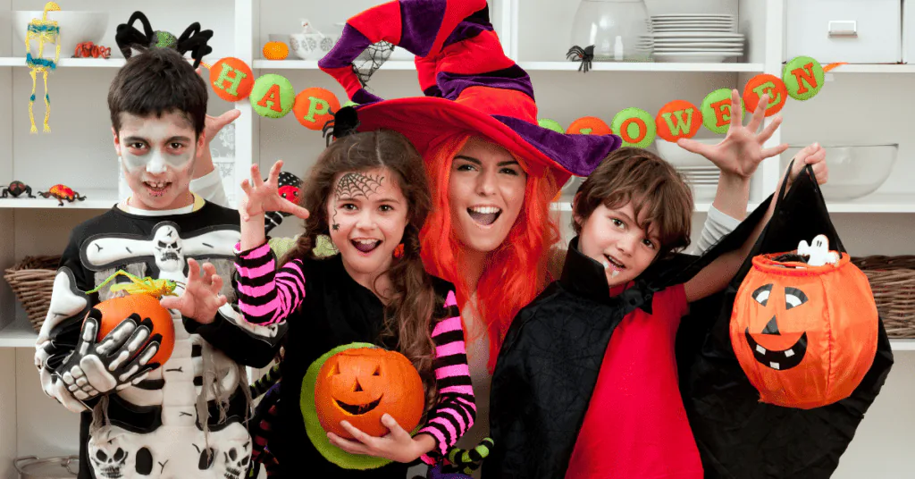 Three children in Halloween costumes and an adult in a witch hat holding pumpkin candy buckets