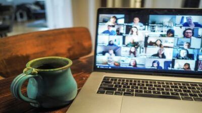 Laptop on a wooden table showing a video call grid of colleagues, beside a green coffee mug