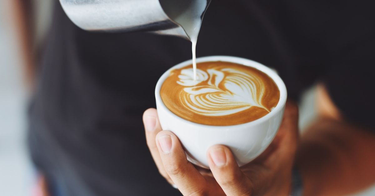 Steamed milk being poured into a white cup to form leaf latte art, held in two hands