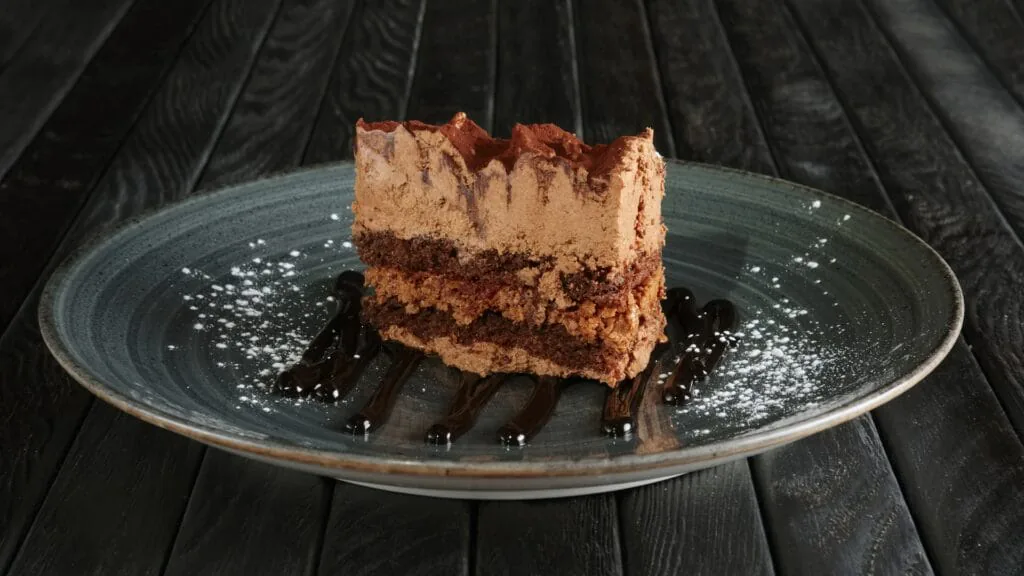 Slice of layered chocolate cream cake dusted with cocoa on a plate dotted with chocolate sauce