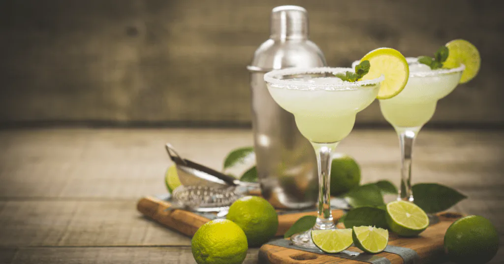 Two salt-rimmed lime margaritas with a cocktail shaker and fresh limes on a rustic wood bar
