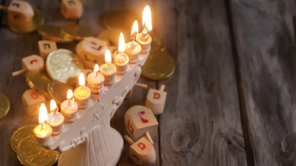 Lit Hanukkah menorah surrounded by dreidels and gold foil chocolate coins on a wooden table