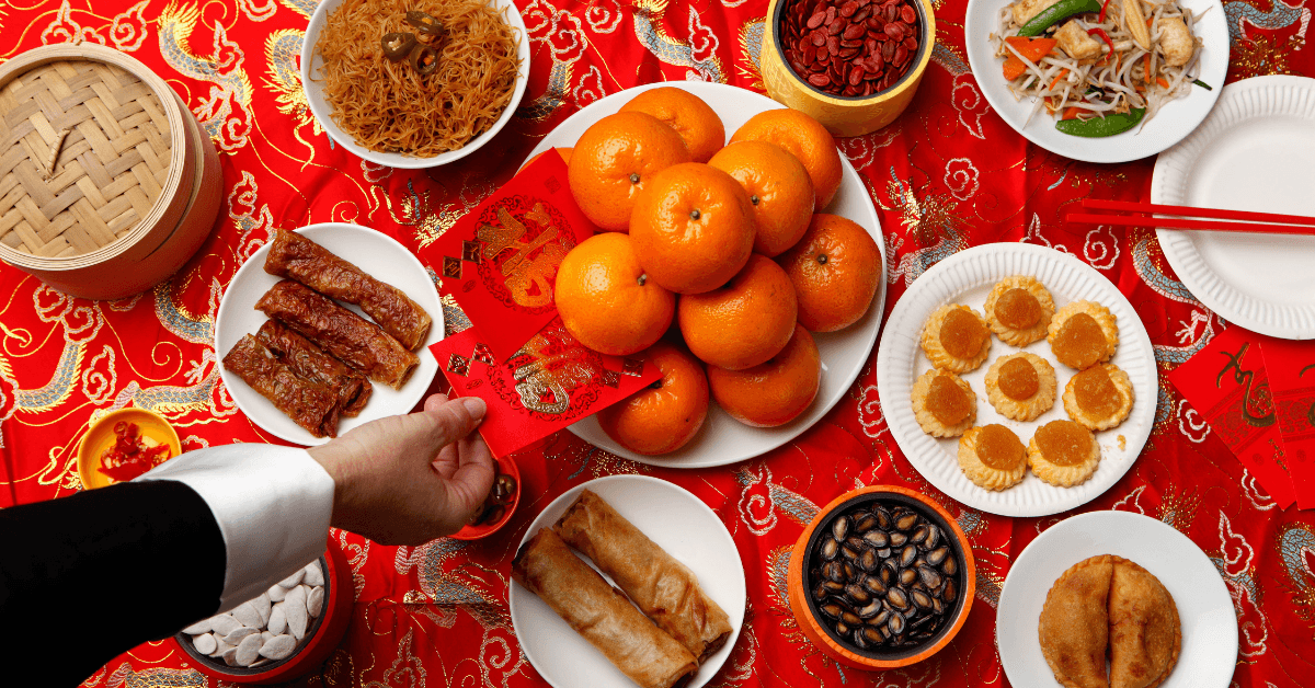 Overhead Lunar New Year spread of mandarins, roast pork and dumplings on a red cloth with a red envelope