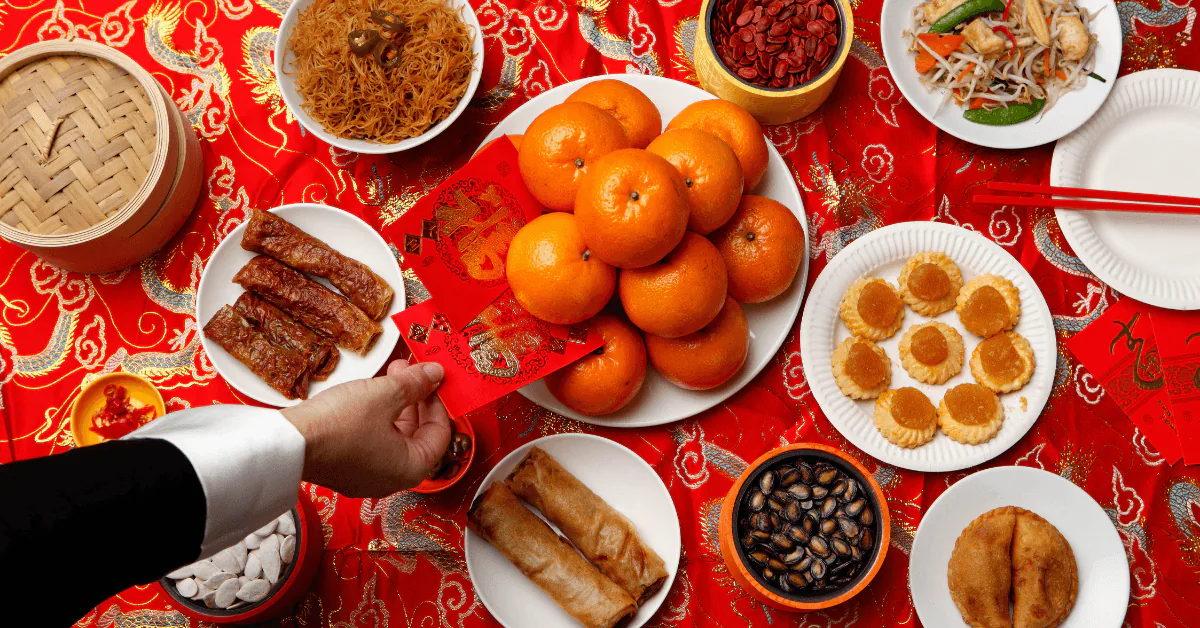 Overhead Lunar New Year spread of mandarins, roast pork and dumplings on a red cloth with a red envelope