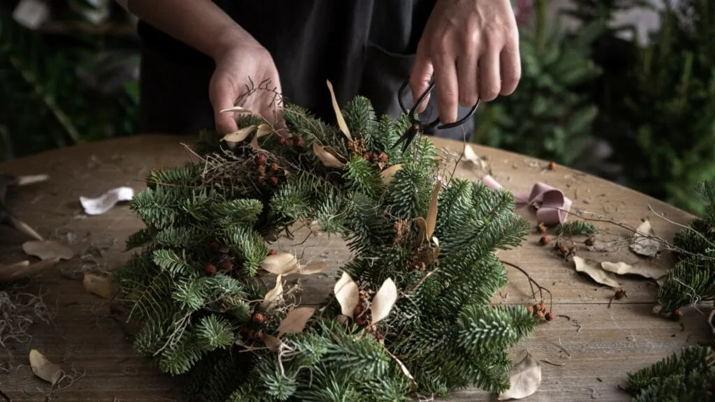 Person assembling an evergreen wreath with dried flowers and shears on a wooden table