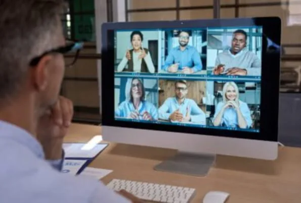 Man at a desk looking at a monitor showing a six-person video call grid