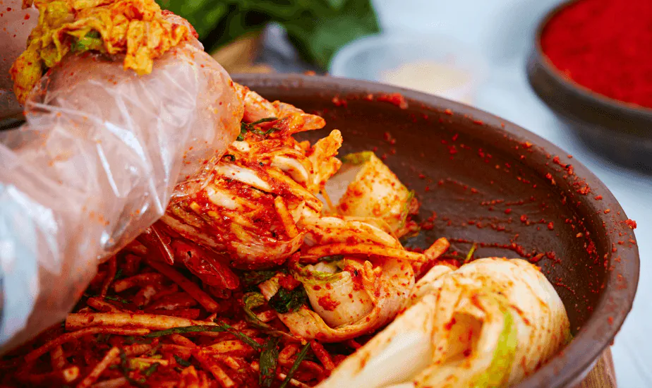 Gloved hands coating napa cabbage in red chili paste in a large earthenware bowl