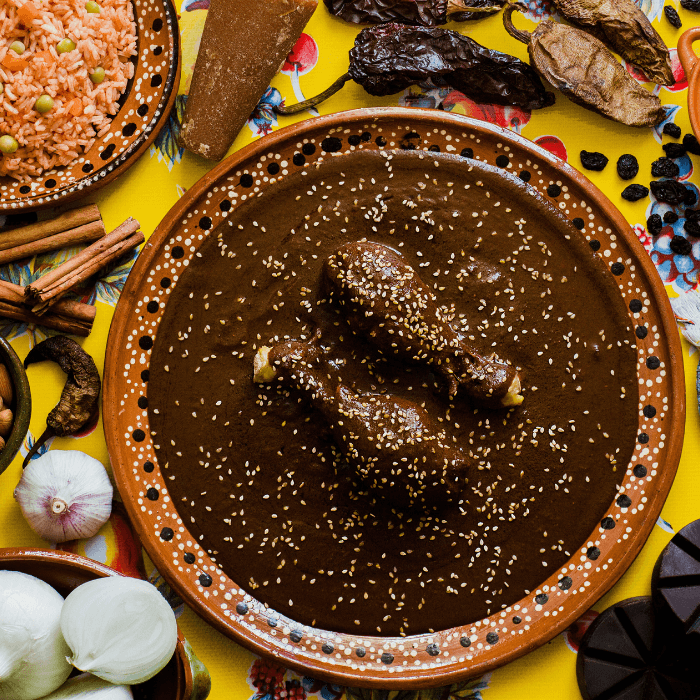 Dark mole sauce topped with sesame seeds in a clay dish, ringed by dried chiles, garlic and rice