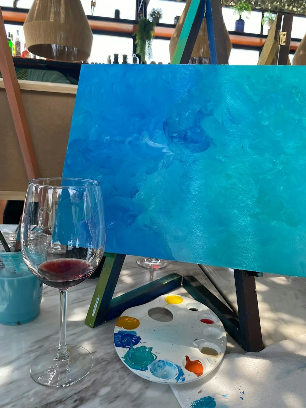 Turquoise ocean painting on an easel beside a glass of red wine and a paint palette