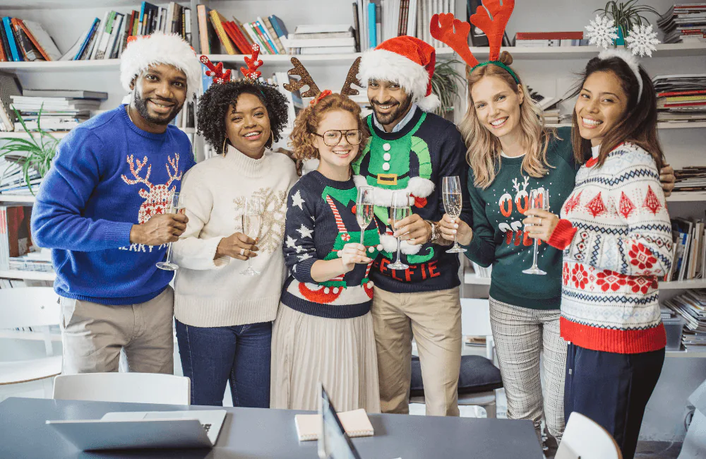 Six coworkers in ugly holiday sweaters and Santa hats toasting with champagne in the office